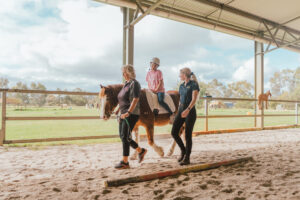 Two women walking at a horse stable with a young girl who is sitting on a horse